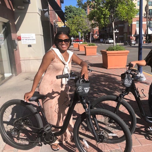 一个人微笑着，站在阳光明媚的城市人行道上，旁边是一辆黑色自行车，背景是大型花坛和建筑物。