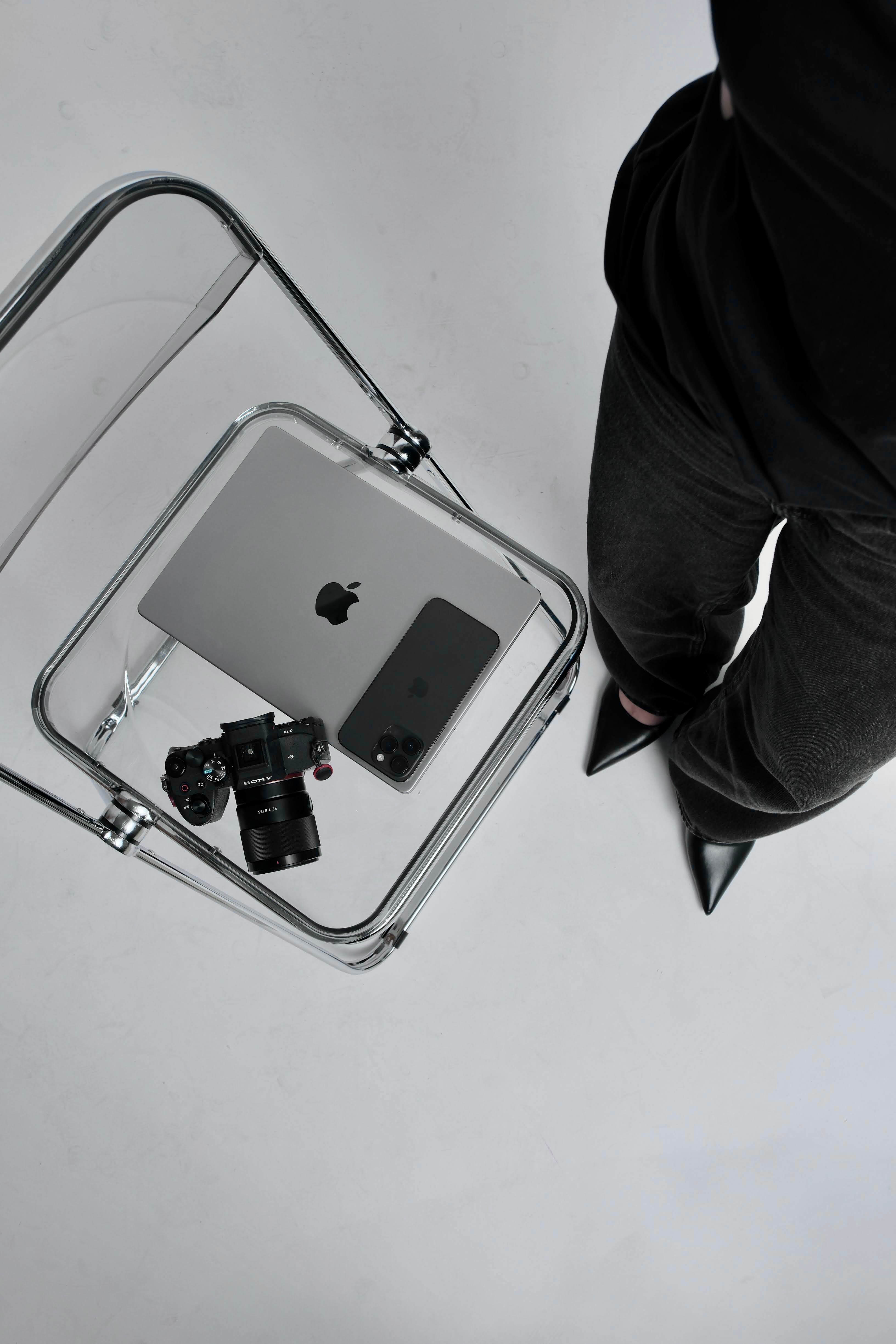 A photo taken from a high vantage point of a woman weating black slacks and black pointy toed stillettos standing next to a clear folding chair with a laptop phone and SLR camera set on top