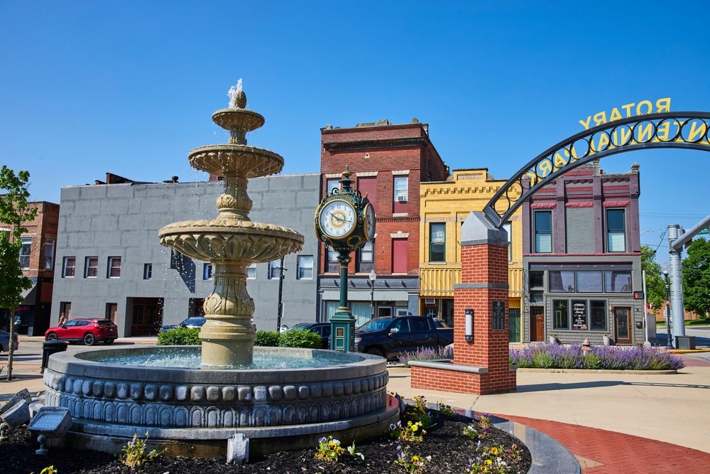 Ornate tiered fountain in downtown Huntington, Indiana, with a vintage street clock, historic brick storefronts, and a Rotary Centennial arch in the background under a clear blue sky.