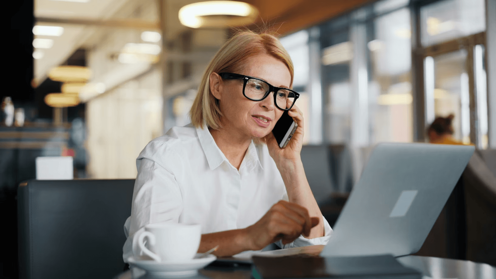A middle-aged woman with glasses and blonde hair talking on a phone while working on a laptop at a café table with a coffee cup nearby