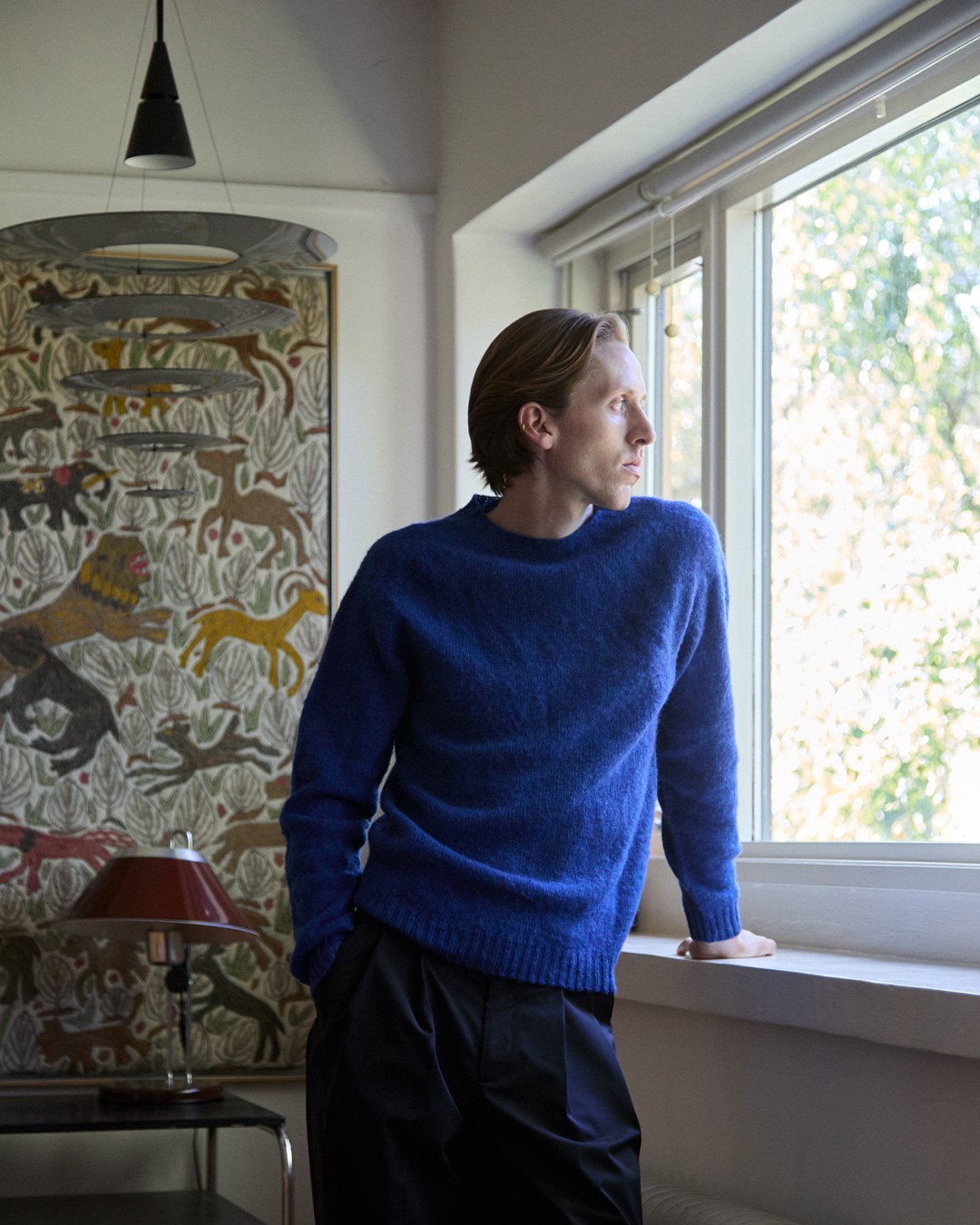 Homme portant un pull bleu roi, regardant par la fenêtre dans le lookbook de Norse Projects