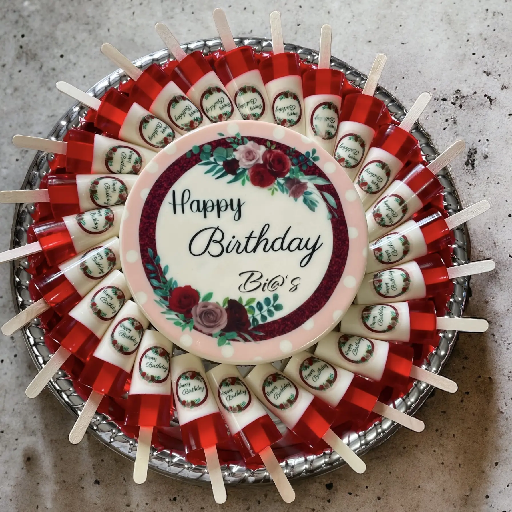 Floral-themed birthday gelatin platter with a central cake featuring “Happy Birthday” text, surrounded by individual gelatin cups adorned with red roses. A sophisticated and decorative treat for a special celebration. Susa’s Sweets & Balloons.