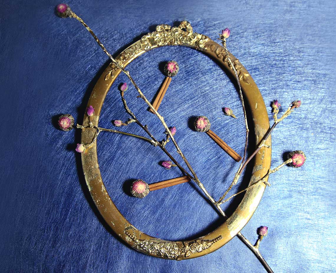 A gold-colored circular frame with ornate detailing rests on a dark blue textured surface. The frame is adorned with small pink flower buds and thin, light brown branches extending outward. Inside the frame are three thin, light brown sticks positioned diagonally. The frame has decorative elements along its edges and appears to have a slightly aged or antique finish. The dark blue background provides a stark contrast to the gold frame and its delicate details.