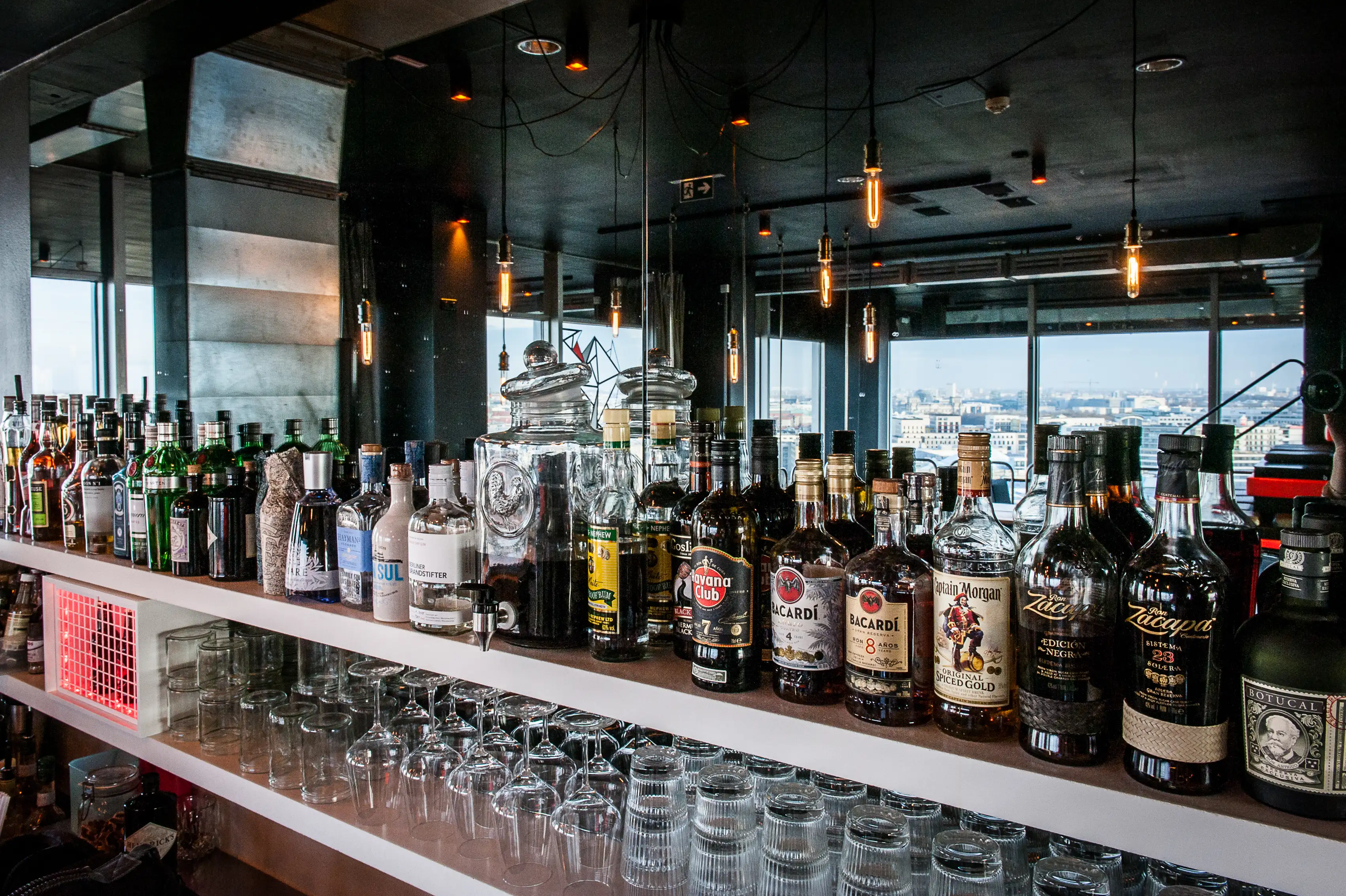 Back bar shelf at SOLAR SKY LOUNGE lined with spirits bottles and stacked glasses, with filament bulbs hanging in front of panoramic windows overlooking Berlin.