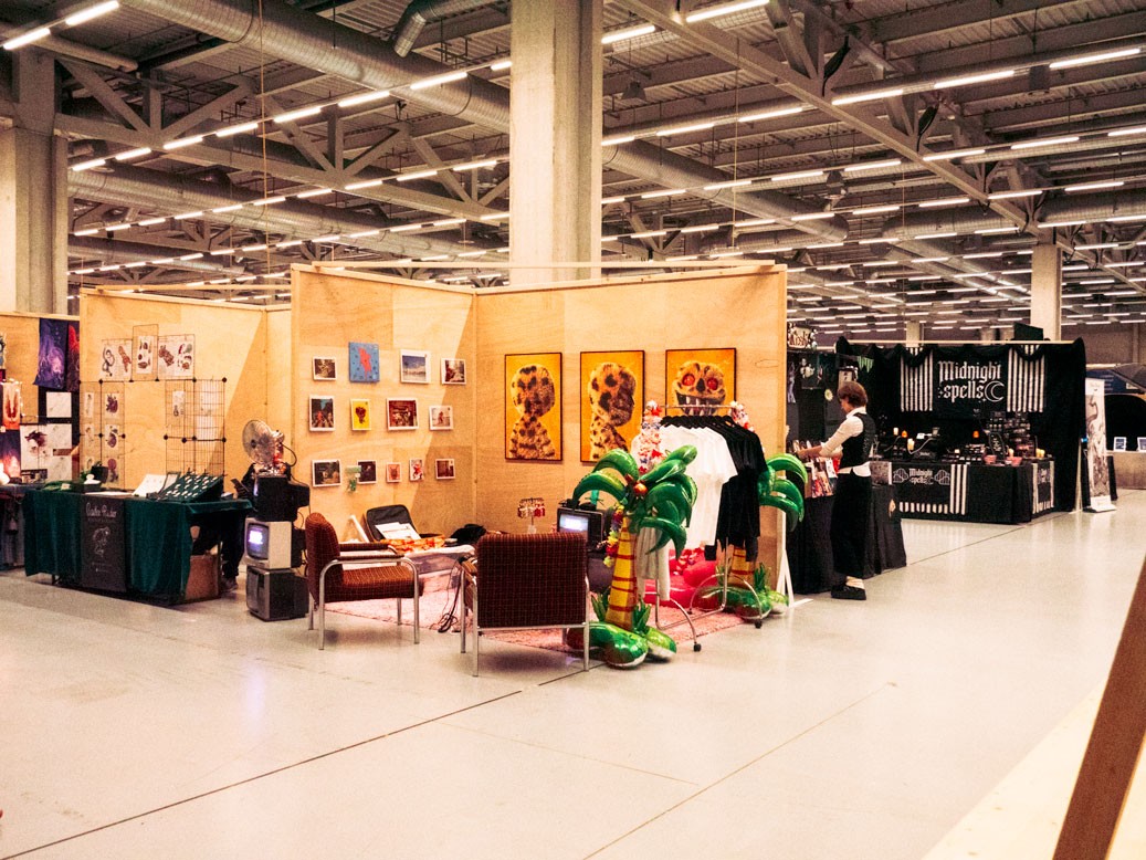 An indoor exhibition space with various artist booths at the Herofest convention in Bern. The booth in the centre of the image is the one of Loveboy and His Imaginary Friends. It features framed artworks, posters, and a small seating area with vintage televisions and inflatable palm trees. Another booth in the background, labeled 'Midnight Spells,' displays gothic-themed merchandise. The space has an industrial ceiling with exposed metal beams and bright overhead lighting, creating a creative and eclectic atmosphere.