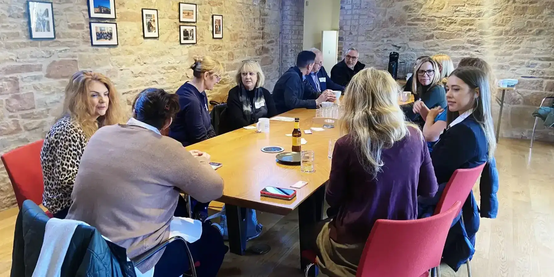 A group of people sit around a wooden table in a cozy, brick-walled room. They are engaged in lively conversation, creating a warm, collaborative atmosphere.