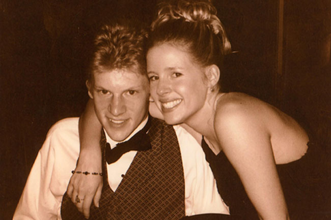 A young man and woman pose for a photograph. The man wears a white shirt, black bow tie, and a patterned vest. The woman wears a dark sleeveless top and has her hair styled up. They stand close together against a dark background. The photograph appears to be in black and white or sepia tone.