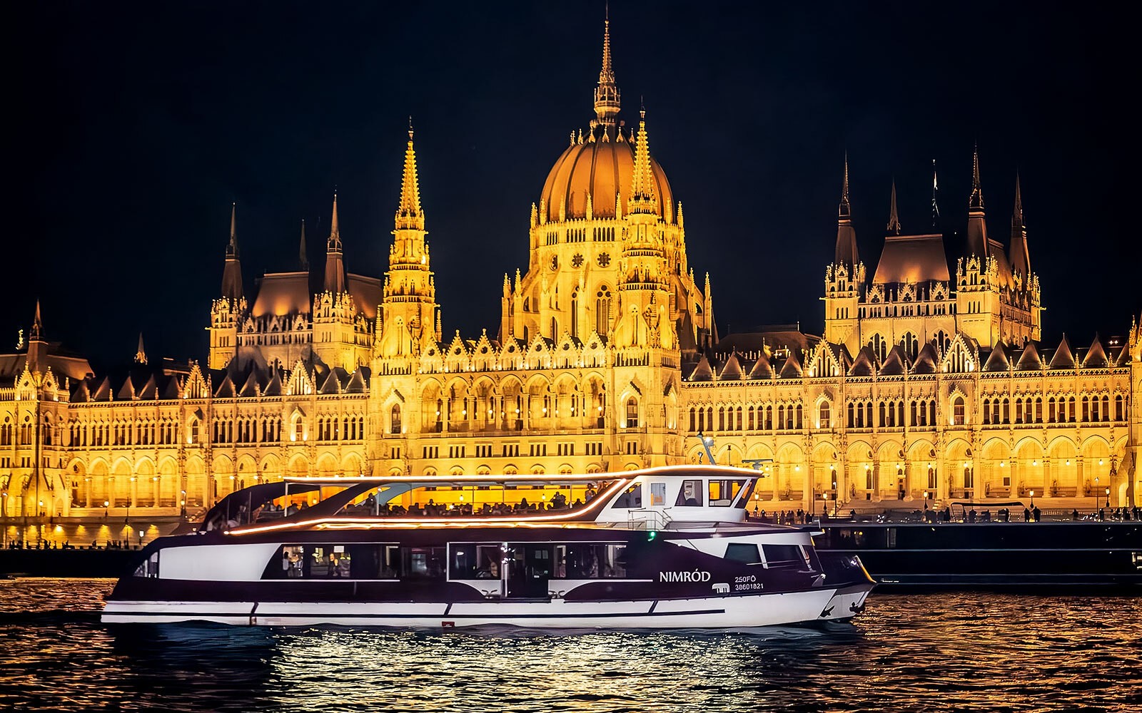Danube river cruise at night with illuminated Budapest skyline and passengers enjoying pizza and beer.