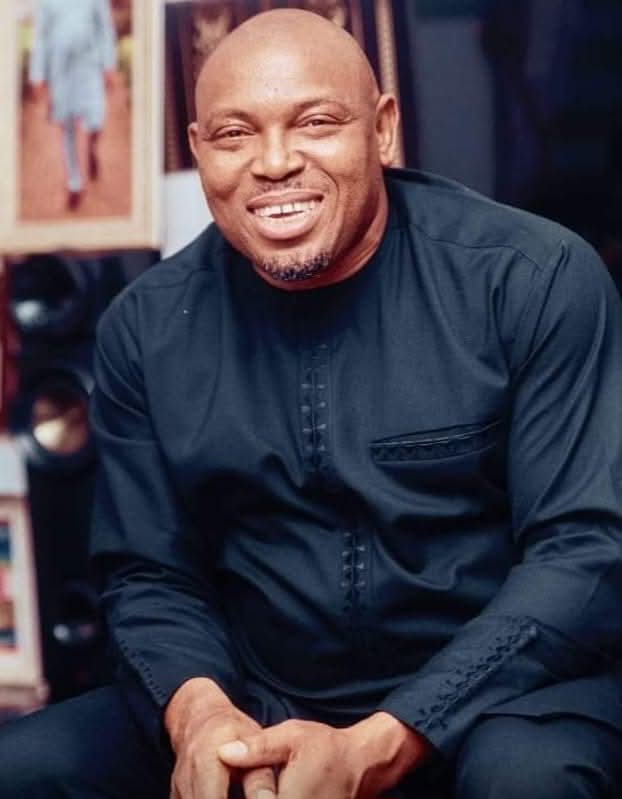Smiling man in a dark suit and tie stands in a well-lit room, exuding confidence and professionalism. Background features blurred office decor.