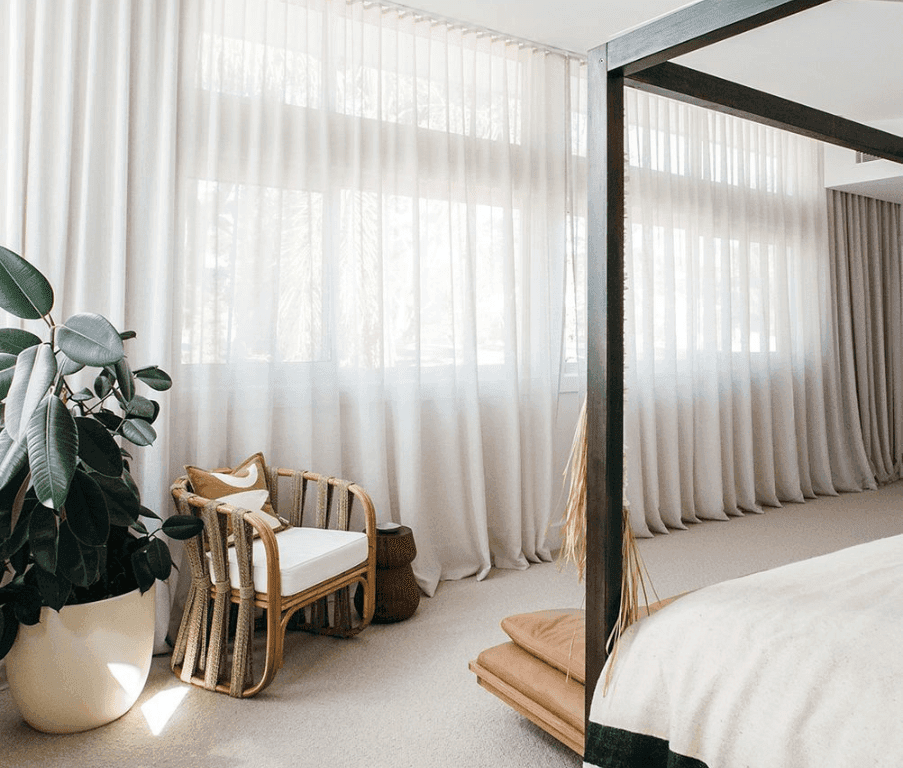 a bright, beautiful, minimalistically designed room, with floor to ceiling drapes, a designer chair, and potted plant at the foot of a bed with frame