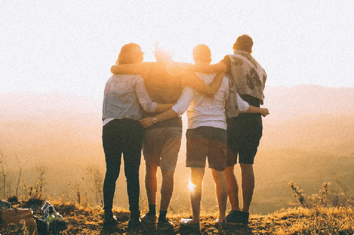 A group of four people are standing close together against a sunlit backdrop, enjoying a moment outdoors.