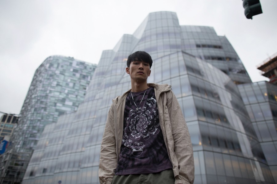 Model wearing graphic T-shirt and jacket stands in front of modern glass building on cloudy day