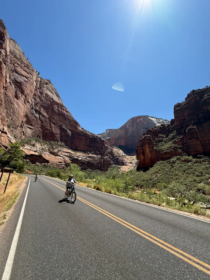 Sara riding downhill on an electric bike with Carrie following close behind on the road inside Zion National Park.
