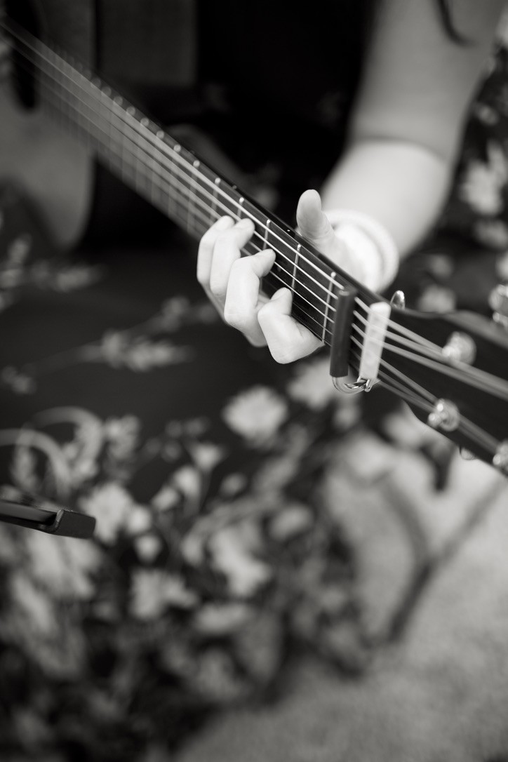 chwarz-weiß-Nahaufnahme einer Hand, die auf dem Griffbrett einer Akustikgitarre Akkorde greift. Ein Kapodaster ist an den Saiten befestigt. Der Hintergrund ist leicht verschwommen und zeigt ein gemustertes Kleid oder Tuch.Maria Wiethaler freie Rednerin und Sängerin für Hochzeiten, Taufen, Trauerfeiern und Events aus München und Landshuter Umgebung