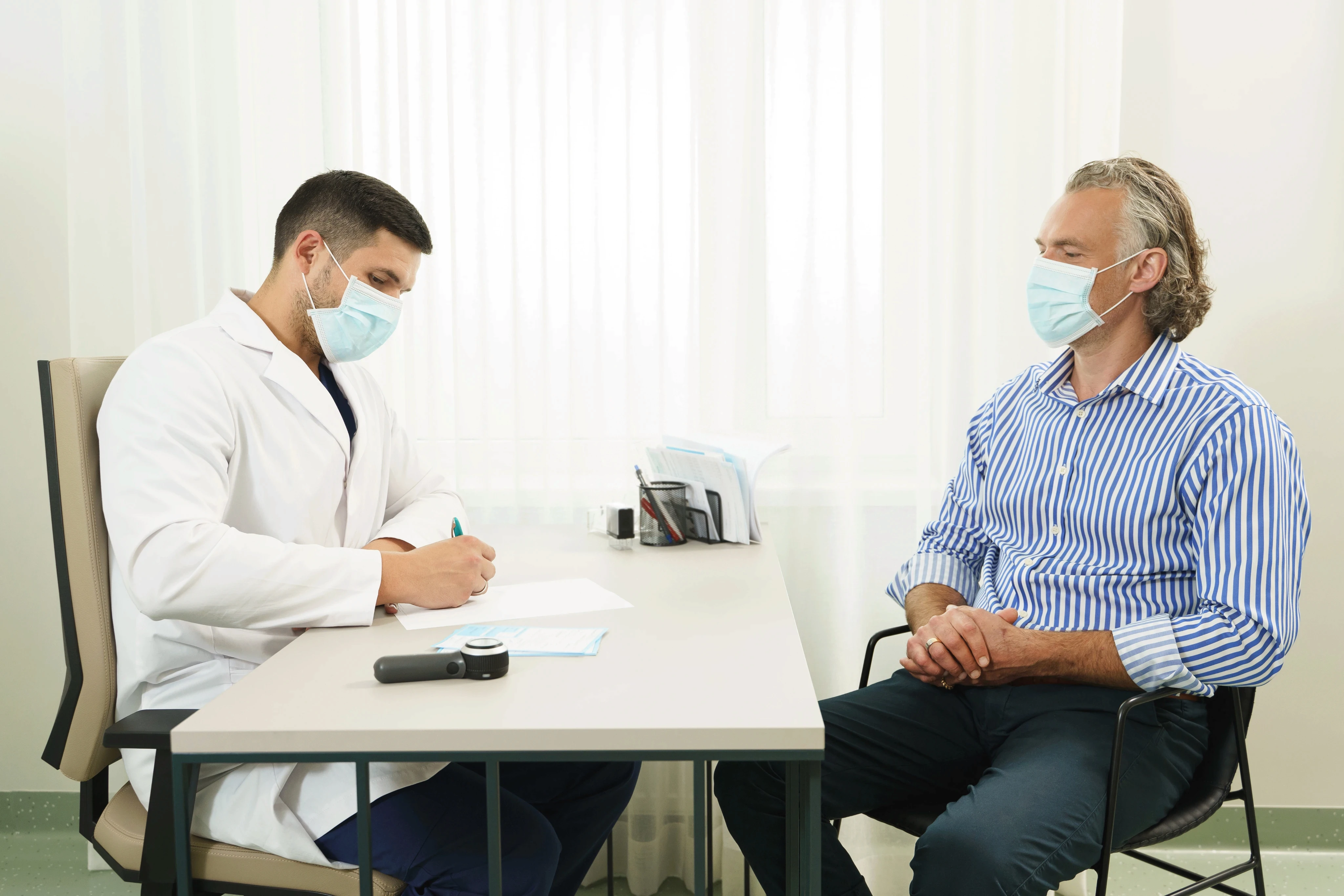 A doctor in a white coat, wearing a mask, writes notes at a desk while a patient in a striped shirt, also masked, attentively listens in a well-lit room.