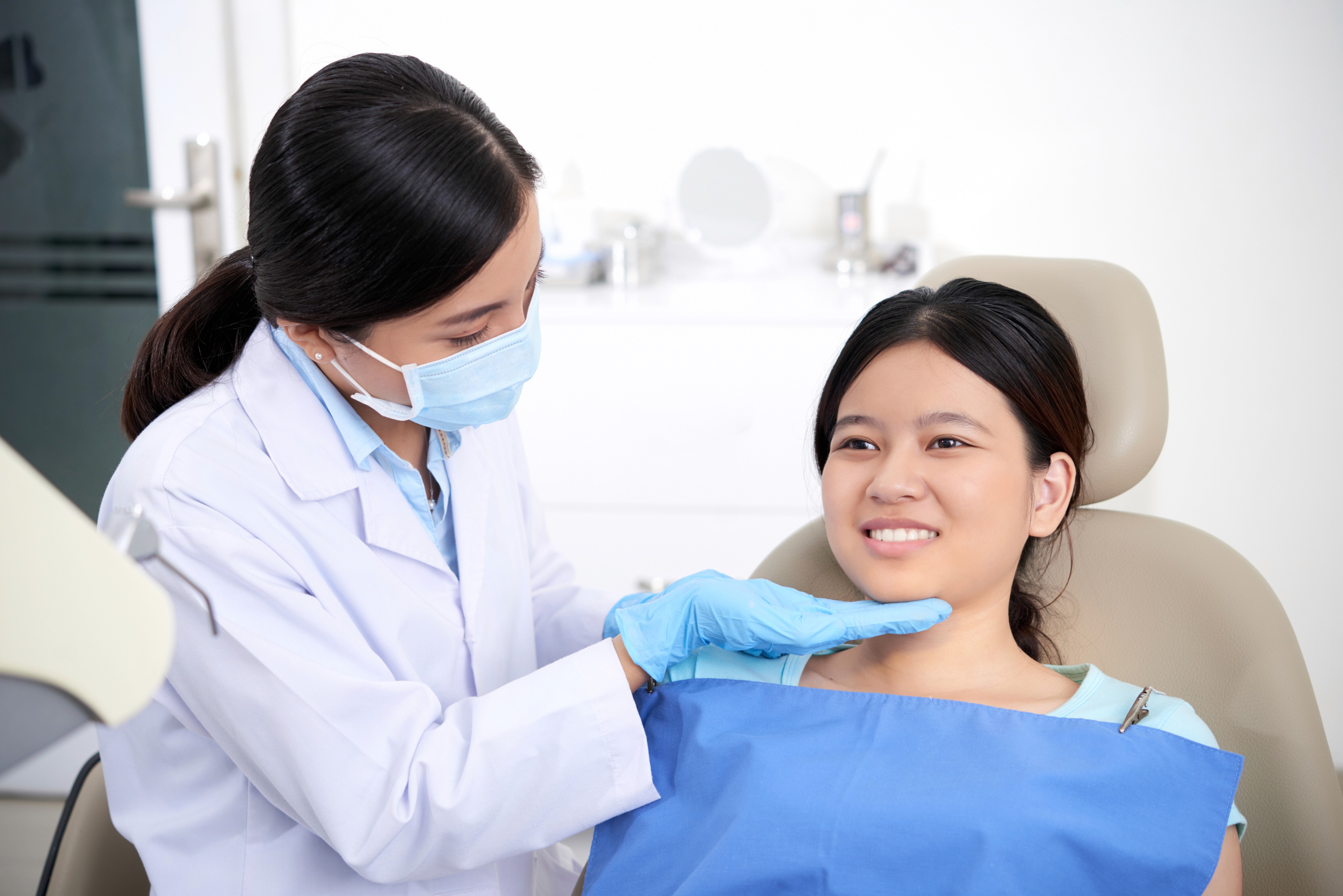 A dental professional wearing a mask and gloves explaining dental care to a smiling female patient sitting in a dental chair