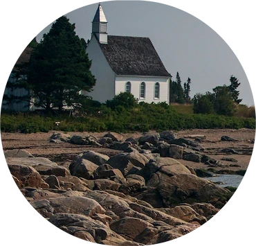 Petite église blanche en bord de mer, entourée de conifères, vue depuis un rivage rocheux sous une lumière douce.