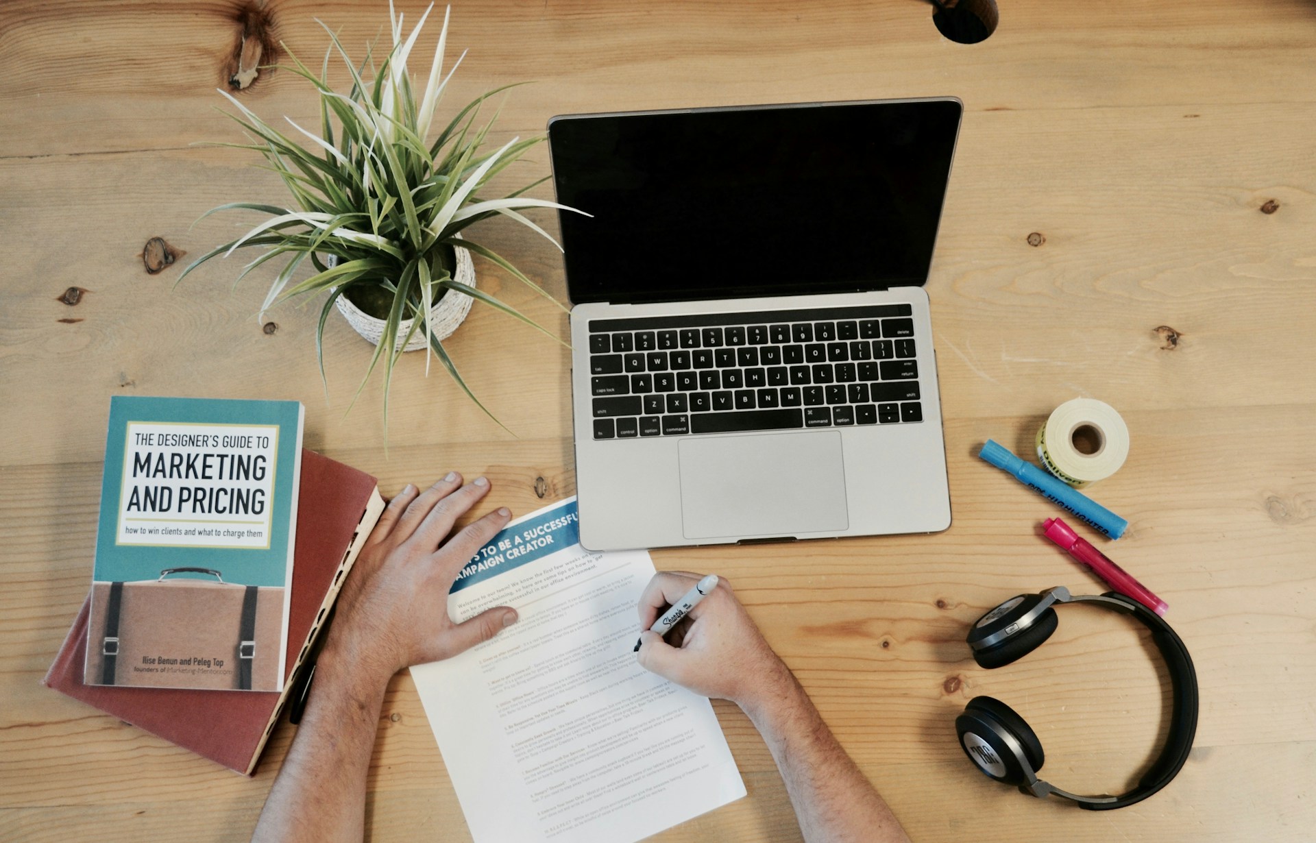 A person sits at a wooden desk, working on a document with a pen. A laptop, headphones, markers, tape, and a potted plant are also on the desk. The person is wearing a white shirt. The document has the words "TO BE A SUCCESSFUL CAMPAIGN CREATE" written on it.