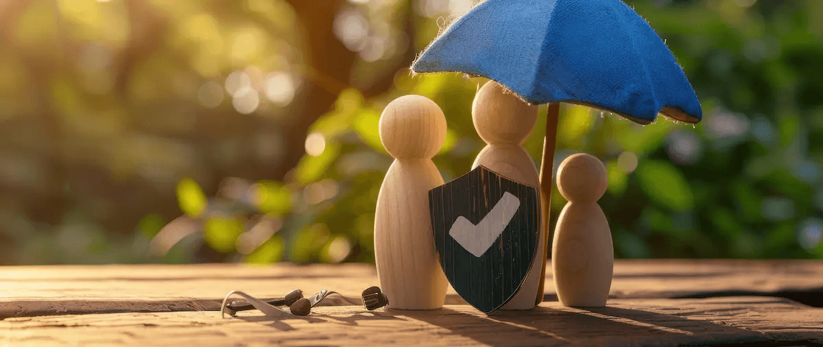 drei Holzfiguren stehen unter einem blauen Schirm - vor ihnen ein Schild mit einem Haken, der für 'Schutz' steht