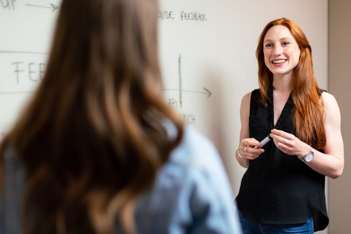 Smiling woman gives a presentation in front of a whiteboard with graphs, speaking to a person facing away from the camera.