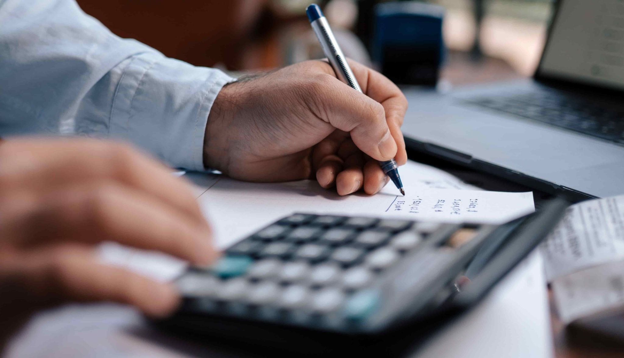Pessoa faz cálculos com calculadora e anota valores em papel ao lado de um notebook.