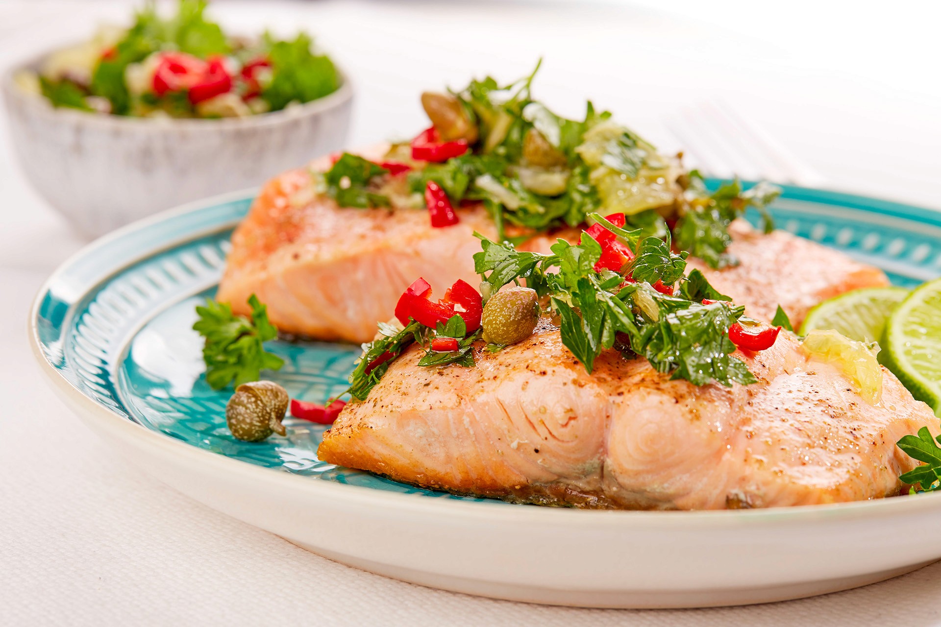 Two grilled salmon fillets garnished with fresh parsley, sliced red peppers, and capers are served on a turquoise plate with lime wedges, accompanied by a small bowl of mixed greens in the background.