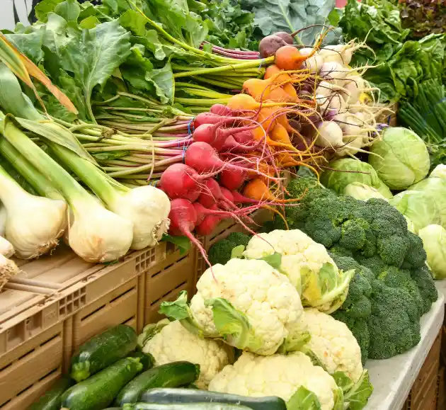 A vibrant assortment of fresh vegetables on display, including green onions, radishes, cauliflower, broccoli, and zucchini, creating a lively market scene.