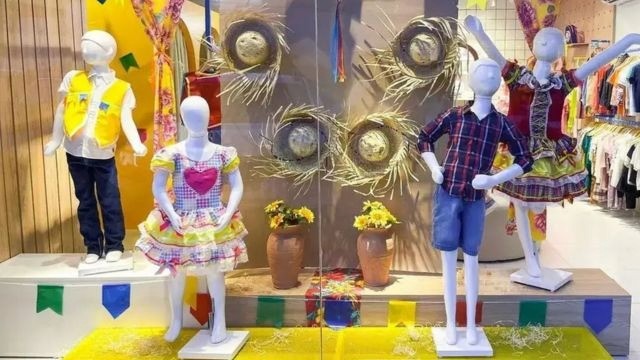 Vitrine com manequins vestindo roupas típicas juninas, chapéus de palha pendurados ao fundo e bandeirinhas coloridas na base.