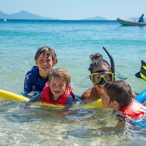 Four children in life vests playing in shallow ocean water, one wearing a snorkel mask. Mountains and a boat are in the background.