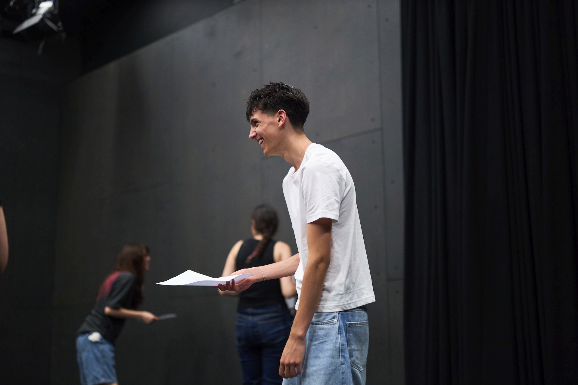 A teenager stands in a rehearsal room holding a script, smiling during a scene exercise with other participants in the background.