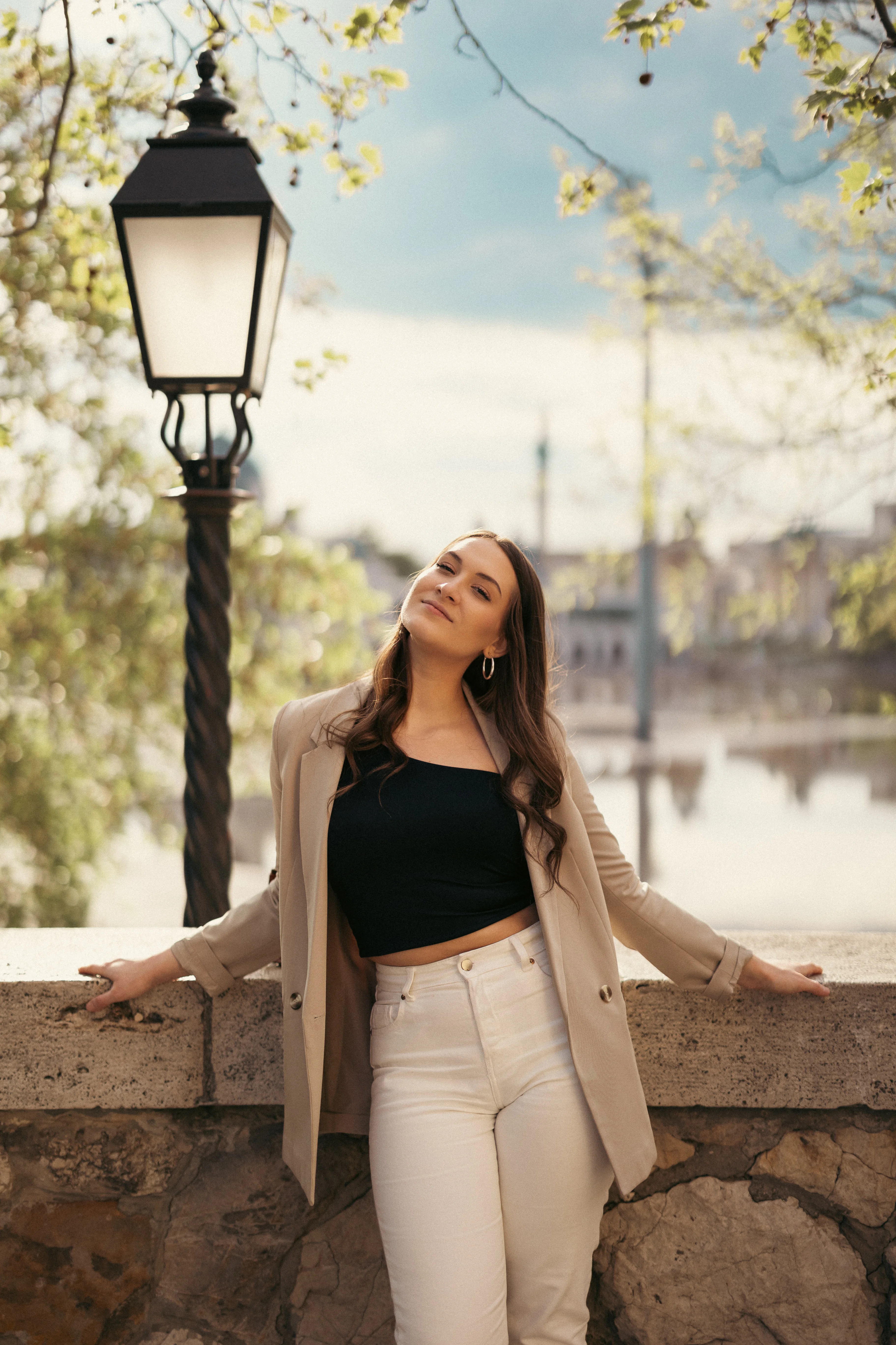 Fiatal nő elegáns casualben támaszkodik kőkorlátnak tavaszi/nyári napfényben, történelmi lámpaoszlop és Városliget tó a háttérben, Vajdahunyad vára közelében