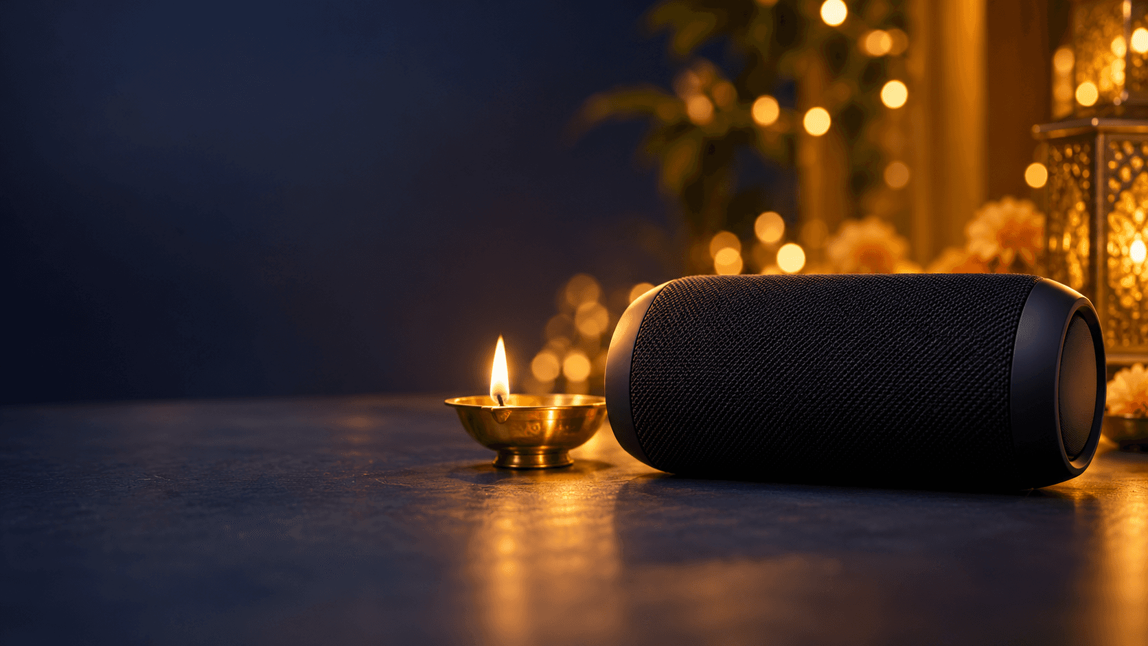 Black wireless speaker beside a lit brass diya on a dark navy surface with warm festive lights behind