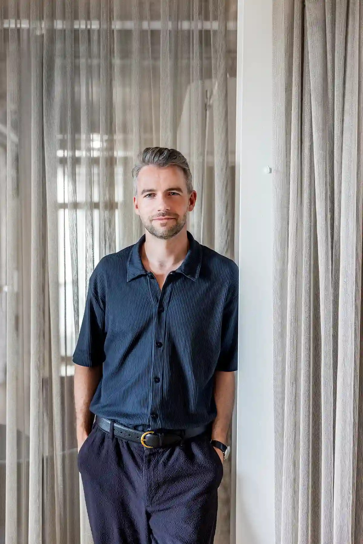 Person mit kurzem Haar und Bart trägt ein dunkelblaues Hemd und steht mit den Händen in den Hosentaschen vor Vorhängen im Büro