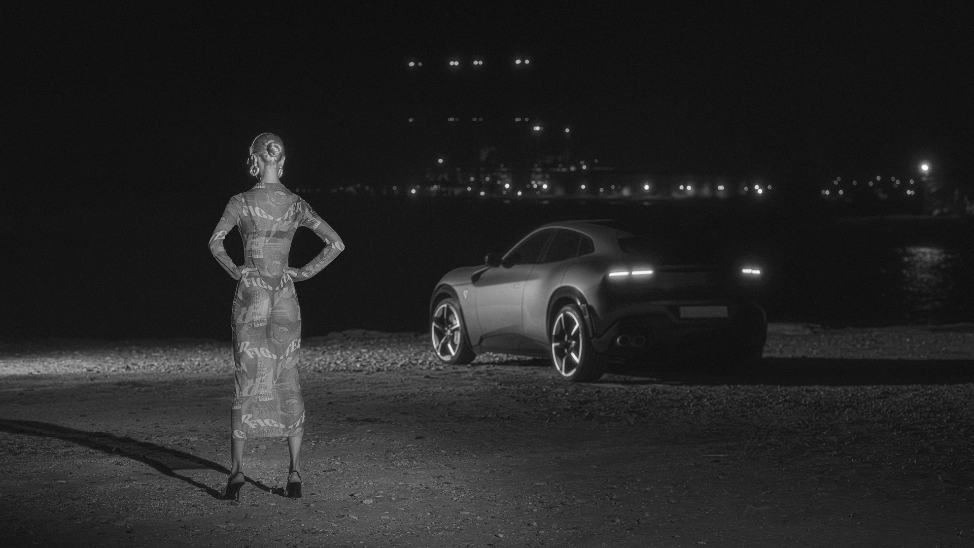 Black-and-white night shot of a model in a printed long-sleeve dress and heels standing with her back to camera, hands on hips, facing a Ferrari Purosangue seen from the rear three-quarter, with harbour lights glittering across the water behind.