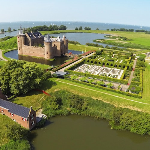 Luftfoto af et slot med en voldgrav, tilstødende haver, en vandkrop i baggrunden og en nærliggende mindre bygning.