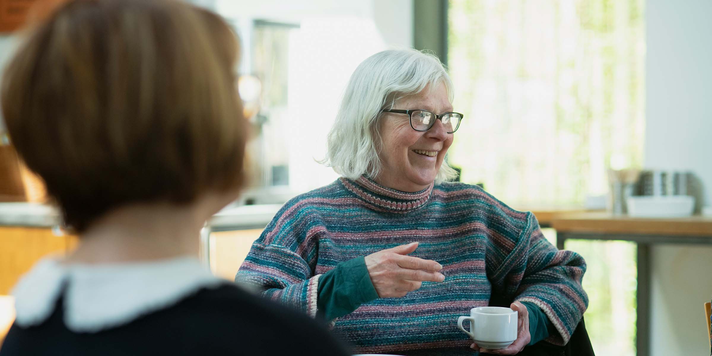 Older lady with white hair and glasses holding a tea cup chatting with other people