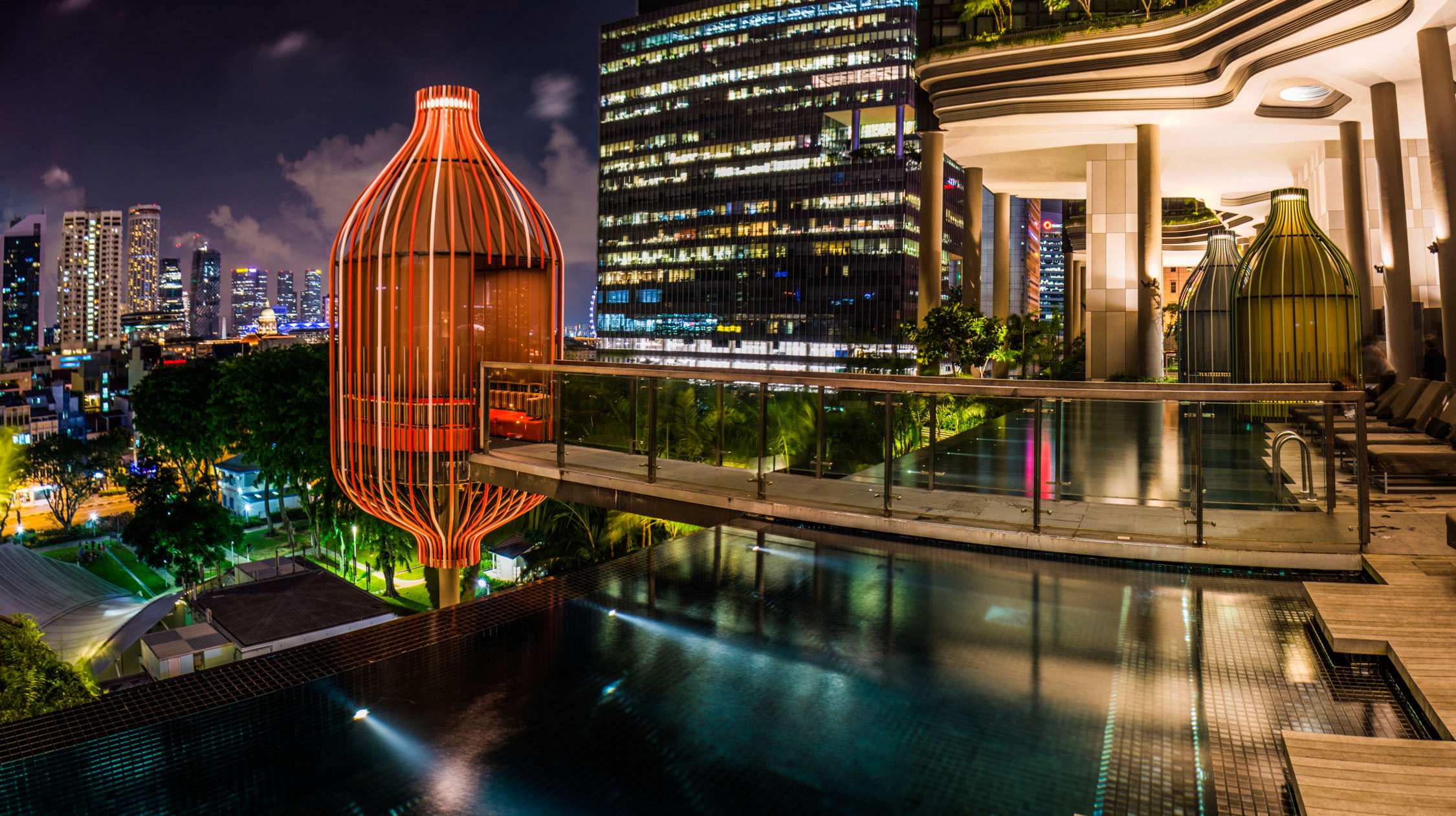 A modern rooftop pool at night features a large orange cylindrical sculpture and a city skyline with lit skyscrapers.