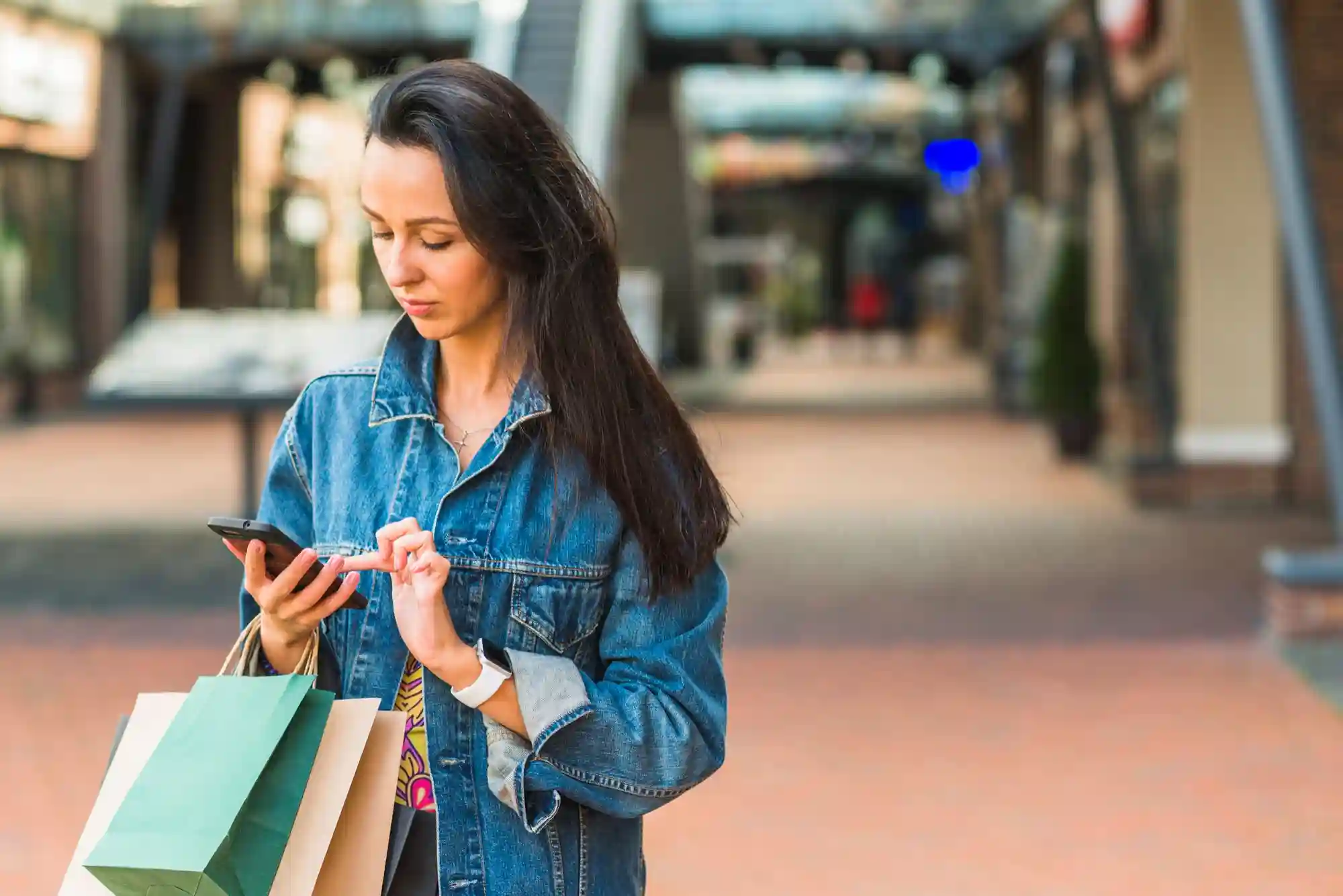 A young woman in a denim jacket checking her phone while holding several shopping bags in a modern outdoor mall.