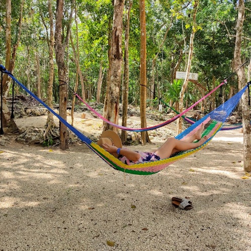 Person relaxing in a colorful hammock in a wooded area, surrounded by trees and other hammocks, with slippers on the ground.