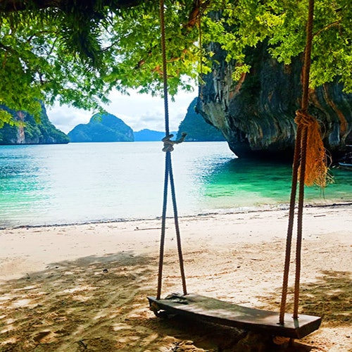 Isang kahoy na duyan na nakasabit sa puno sa mabuhanging dalampasigan na tanaw ang malinaw na asul na tubig at mga likod ng batong apog sa likuran.