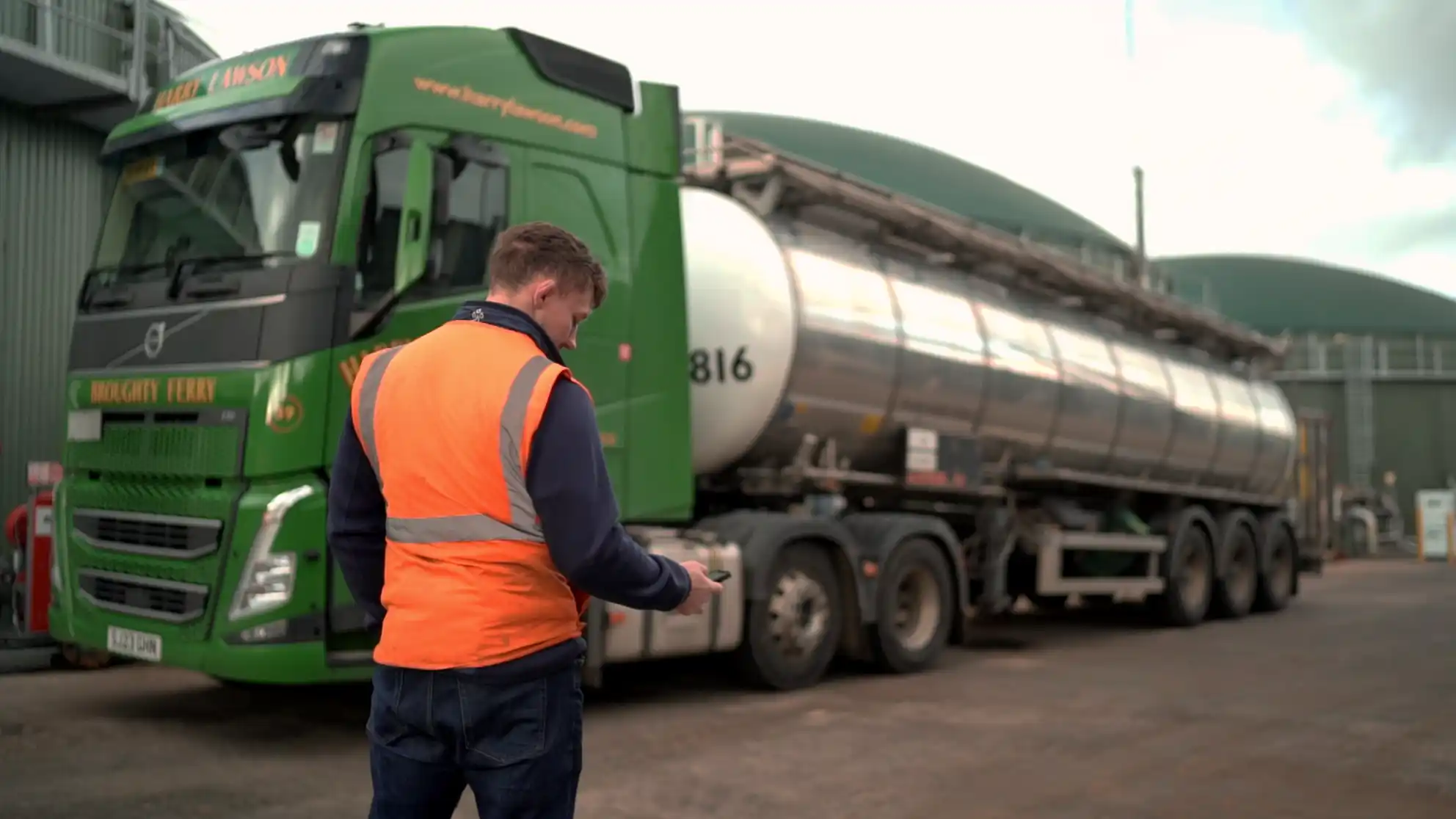 A man in an orange safety vest stands in front of a large green and silver tanker truck, using a smartphone. The scene is industrial and focused.