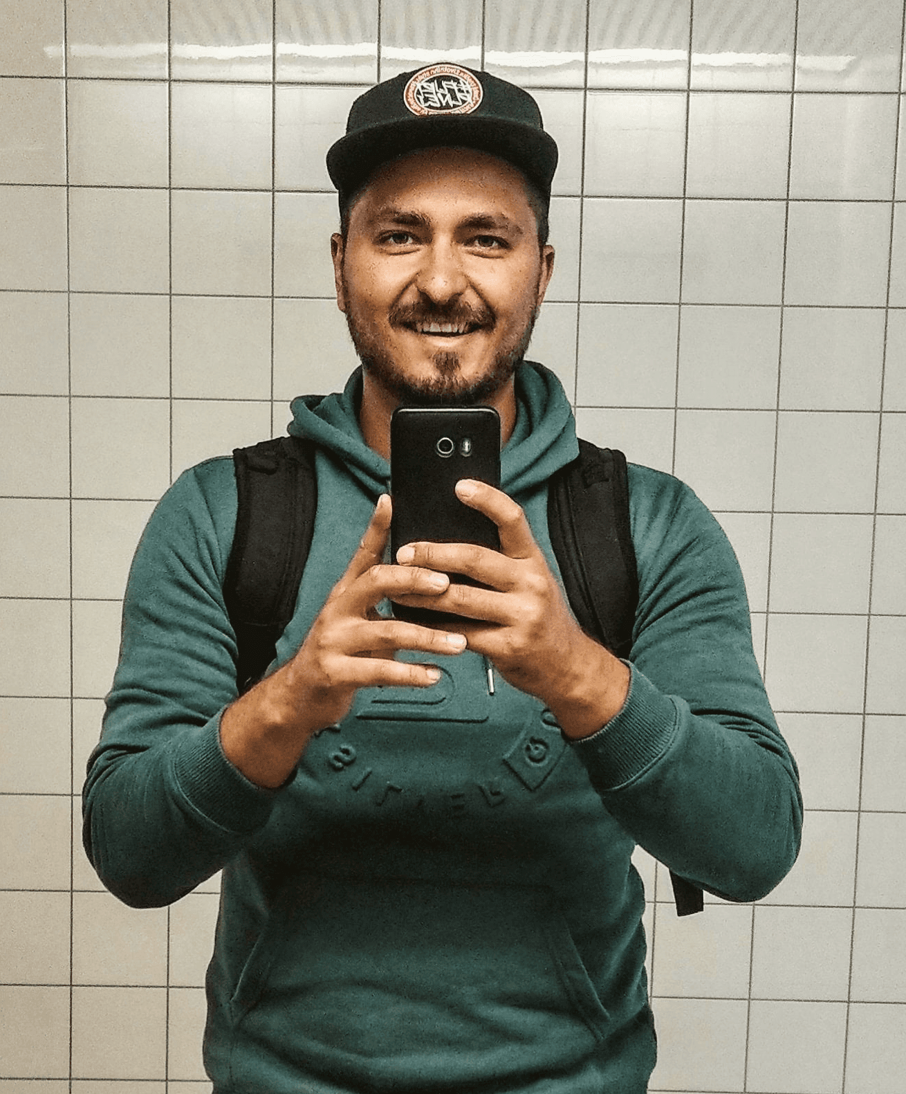 Smiling young man with curly hair, wearing a dark hoodie, against a softly lit background.