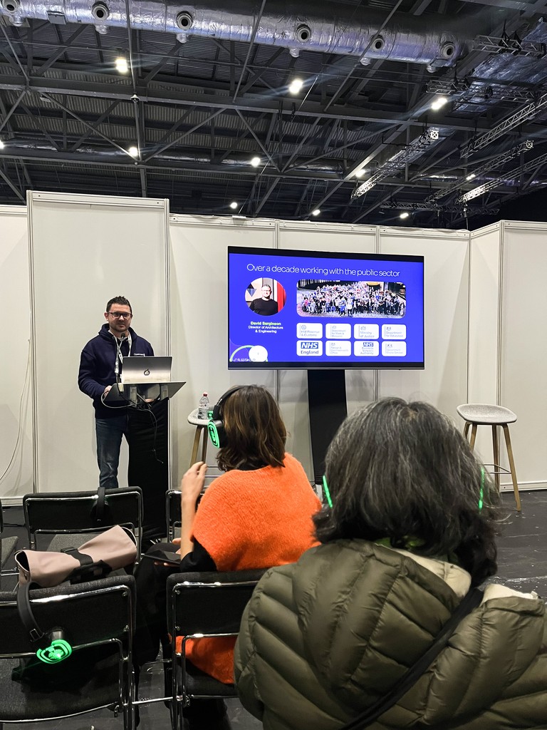 A presenter stands at a podium in a conference booth area, speaking to a small seated audience. A large screen beside him displays a slide about his decade of work in the public sector. Audience members wear green‑lit wireless headsets, and the venue’s high industrial ceiling is visible above the white partition walls.