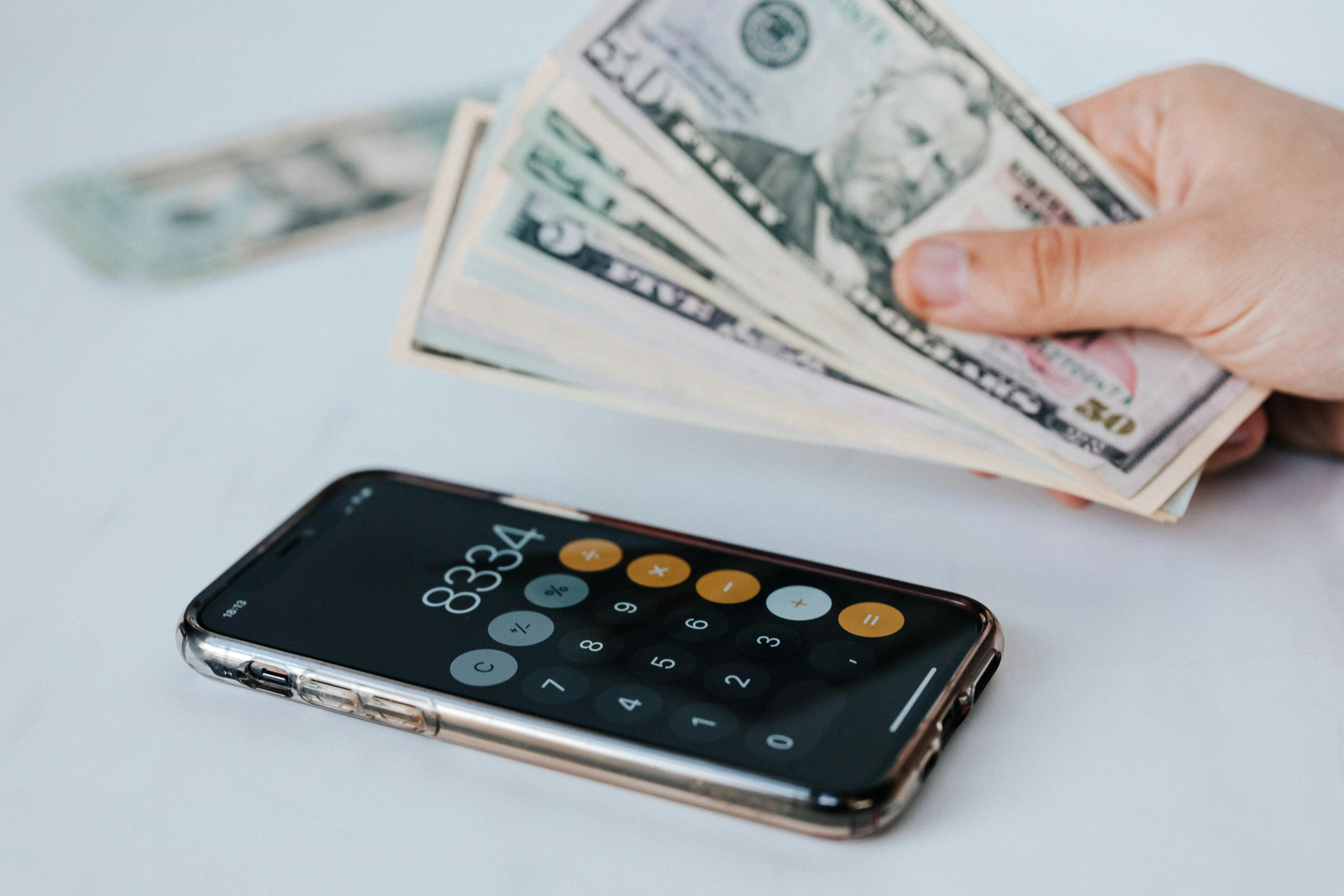 A hand holds cash while a smartphone displays a calculator app on a white surface.