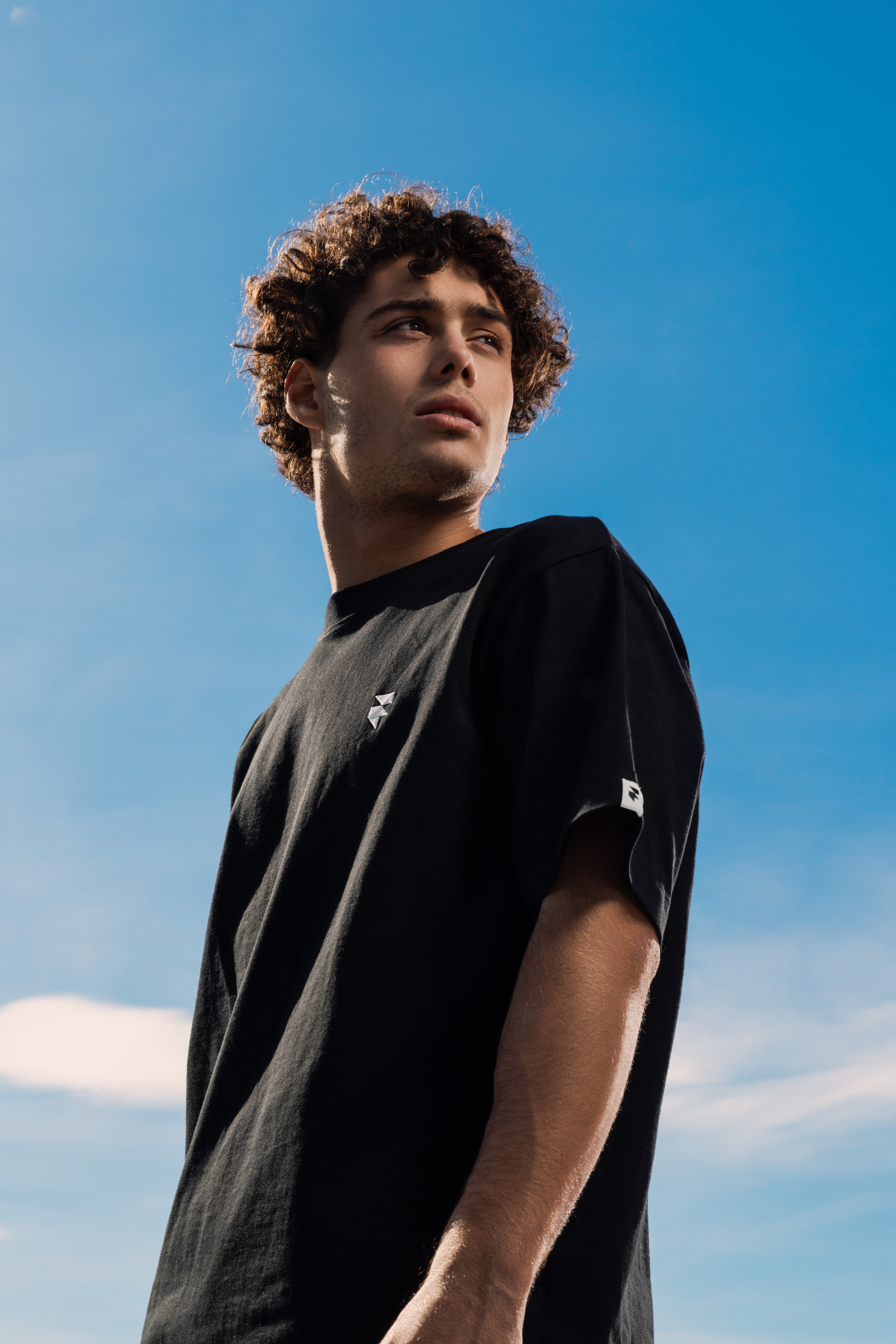 A person with curly hair gazes into the distance against a backdrop of a clear blue sky, wearing a black t-shirt, creating a serene and contemplative atmosphere.
