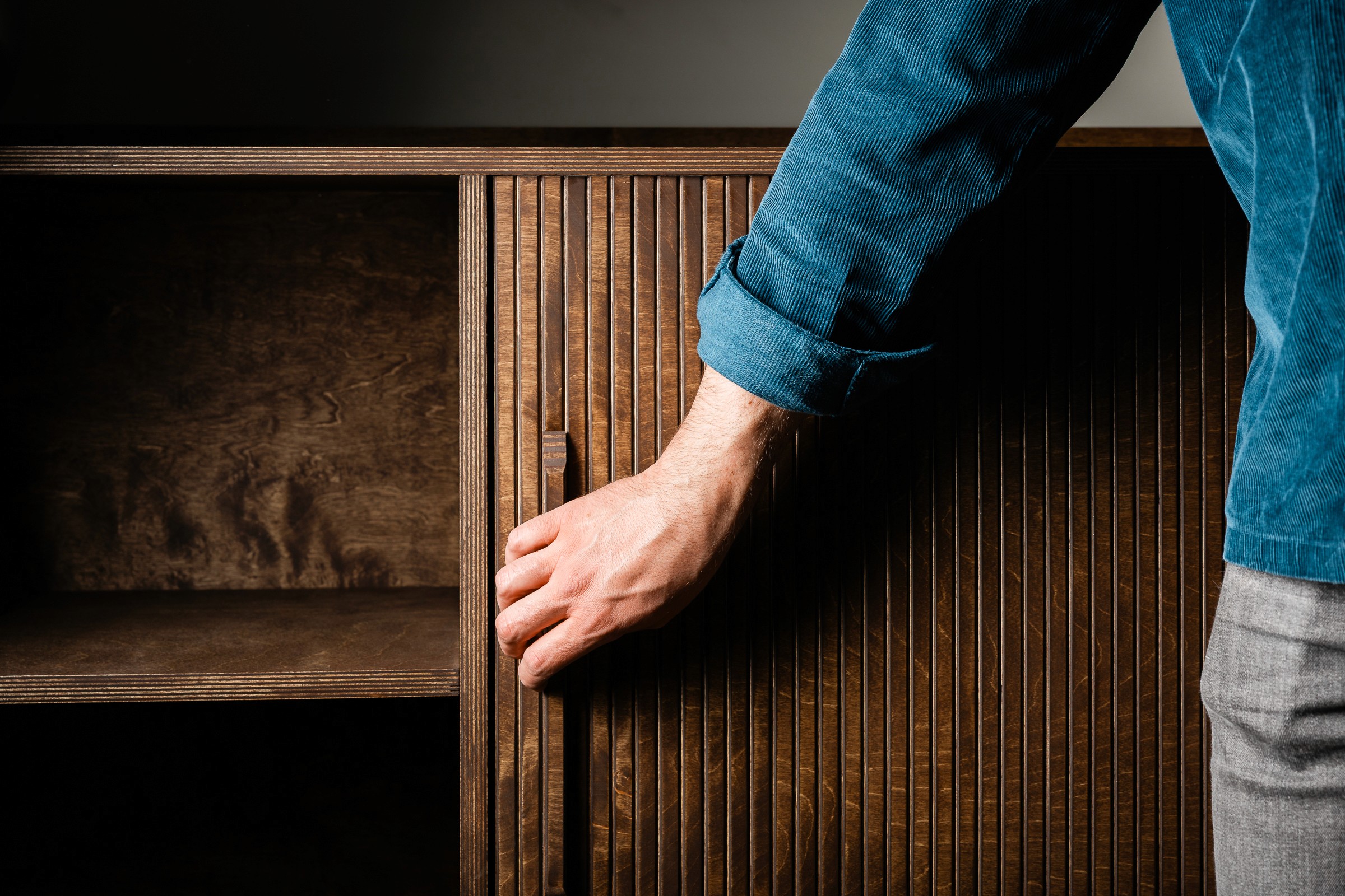 iemand met een grijze spijkerbroek en een blauwgroen geribd overhemd heeft zijn hand op de handgreep van een roldeur van een met de hand gemaakt tv-meuel waar de tv kan worden verborgen