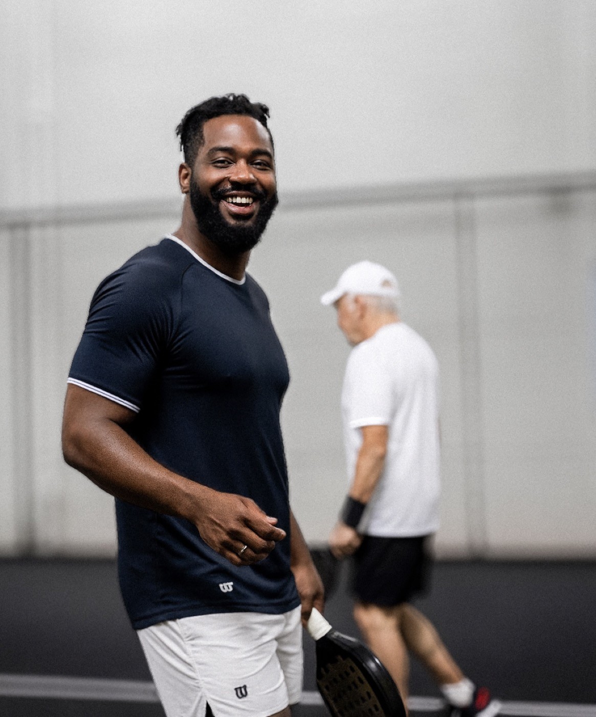 Lachender Padelspieler mit Schläger in der Hand auf dem Court, im Hintergrund ein weiterer Spieler – sportliche Atmosphäre und Freude am Spiel bei Padelnero.