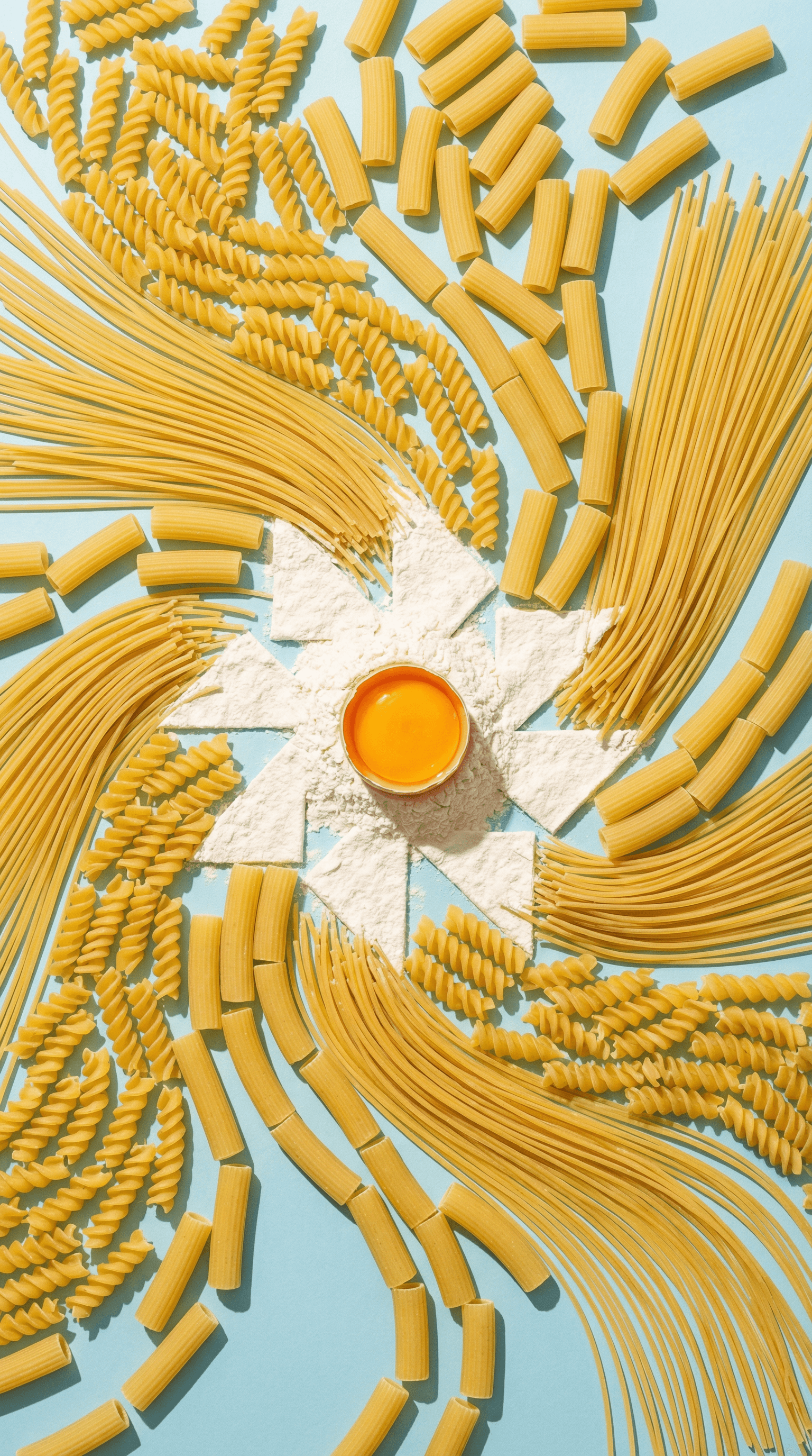 Uncooked pasta shapes arranged in a swirling pattern around flour and an egg yolk.