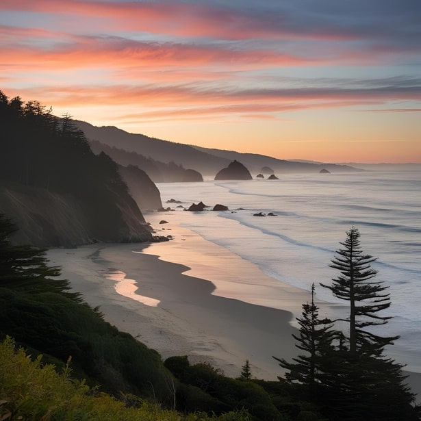 Coastal landscape at sunset with rocky shoreline, sandy beach, ocean waves, silhouetted trees, and misty hills in the distance.