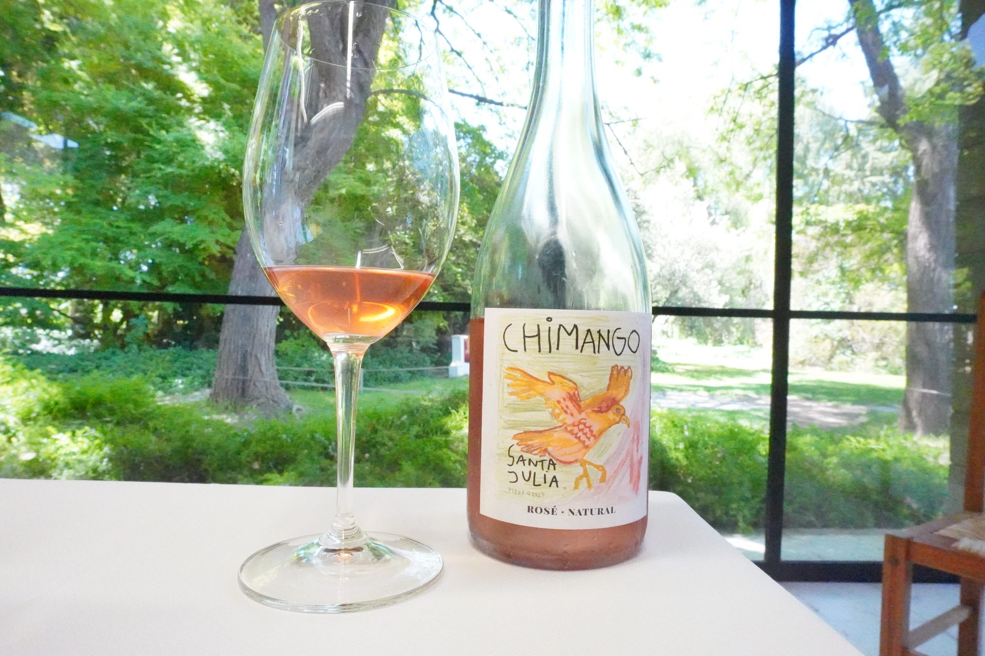 A bottle of Santa Julia Chimango natural rosé beside a glass of rosé on a white table, with green trees visible through the windows.